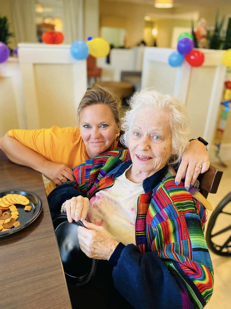 A heartwarming moment captured! A woman and an elderly lady, both wearing bright colors, share a table, celebrating a birthday with balloons and smiles.
