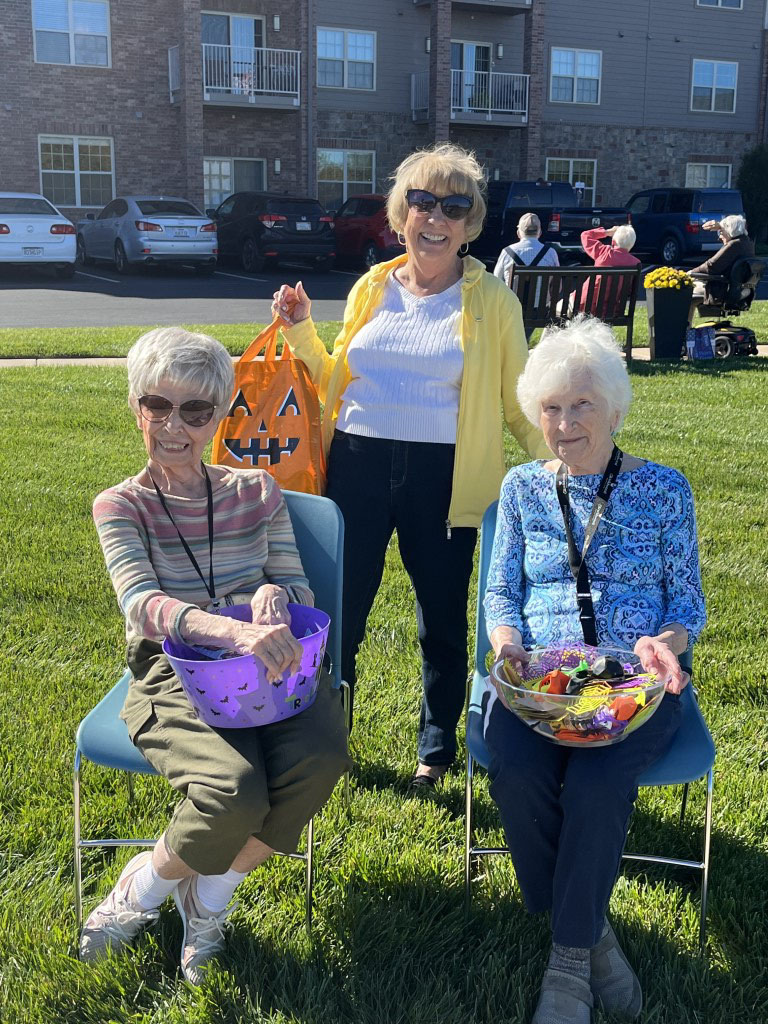 Three senior women with baskets of candy, smiling outside on a sunny day, ready to give out treats to trick or treaters.