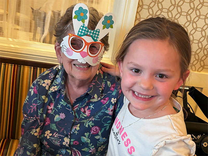 An elderly woman wearing an easter bunny paper mask smiles joyfully with a little girl.