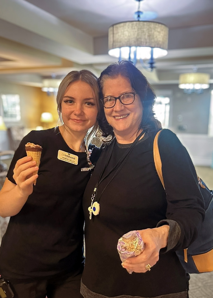 Two women smile and pose together indoors. One holds an ice cream cone, and the other holds a dessert. They are dressed casually, standing in a well-lit room.