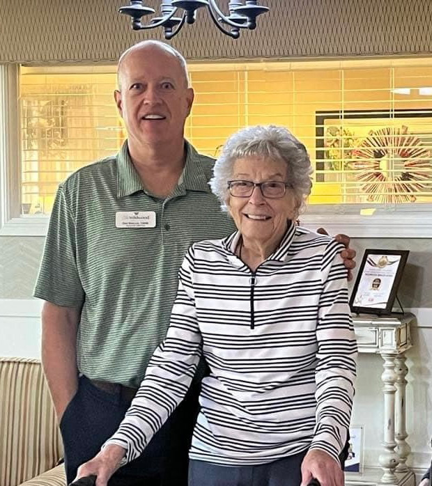 A senior resident stands with a staff member in a senior living community. Both smile at the camera, with the senior resident using a walker and dressed in a striped shirt.