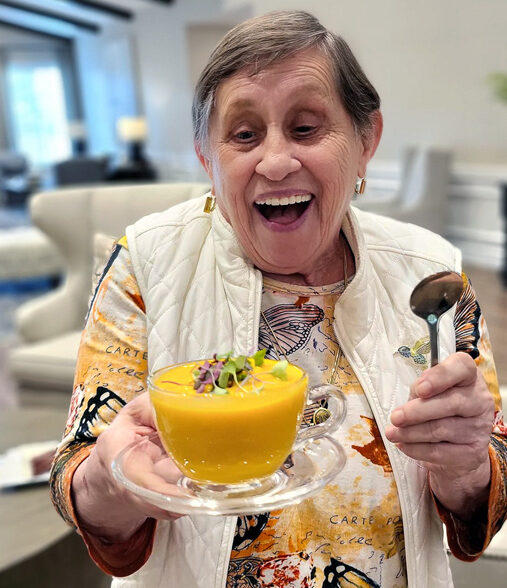 Smiling senior resident joyfully holds a bowl of vibrant orange soup garnished with greens, wearing a colorful butterfly-patterned shirt and white vest, ready to enjoy the meal.