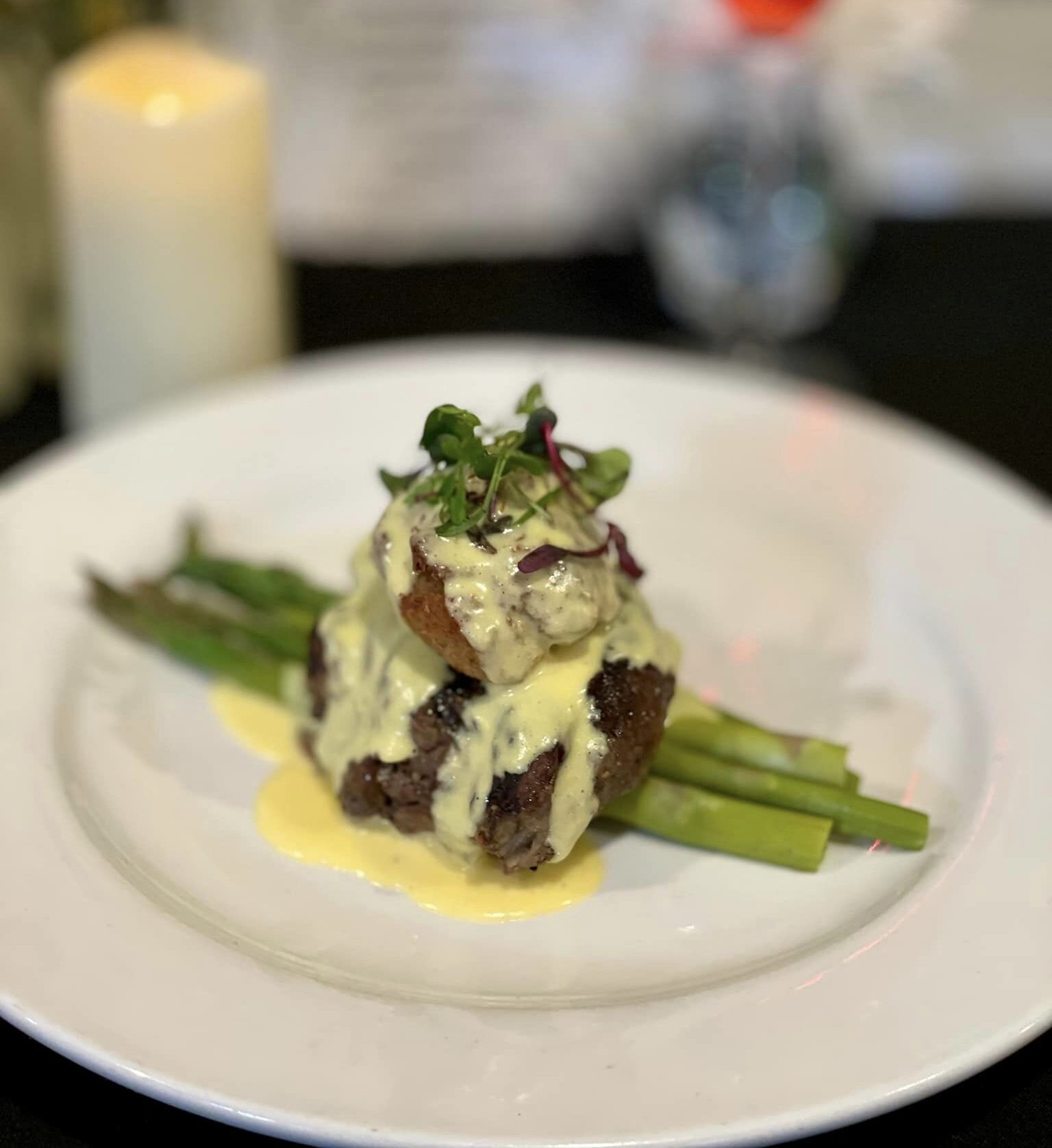 Plated gourmet steak with asparagus, topped with creamy hollandaise sauce and garnished with microgreens, set against a blurred background of a candle, flowers, and drink.