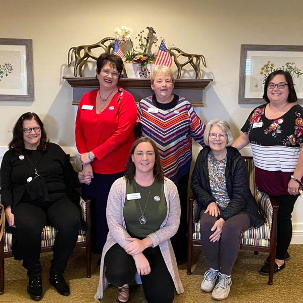 A group photo of senior residents and team members in a senior living community, smiling and posing together in front of a decorated mantel with patriotic elements.