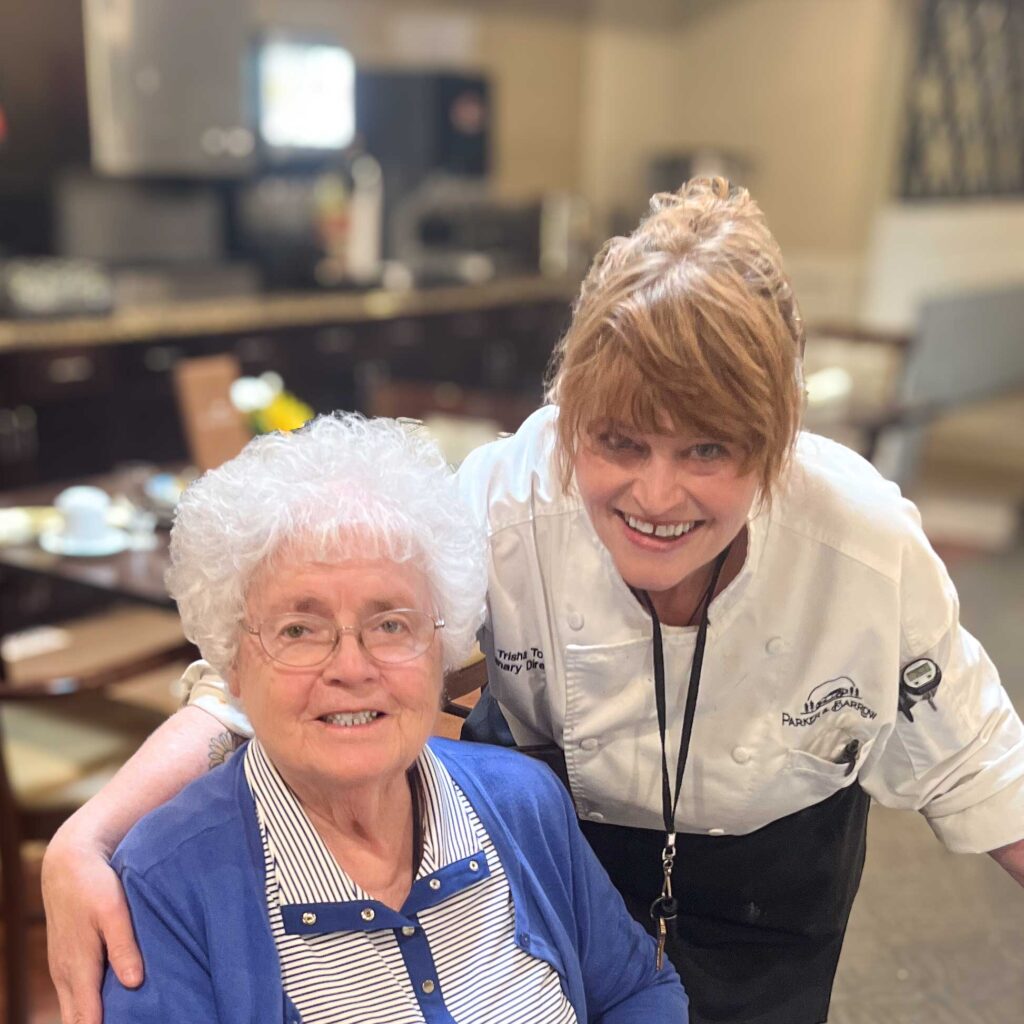 Culinary Director in a chef coat smiles beside a senior resident in a blue sweater at a dining table, highlighting warm hospitality at the senior living community.