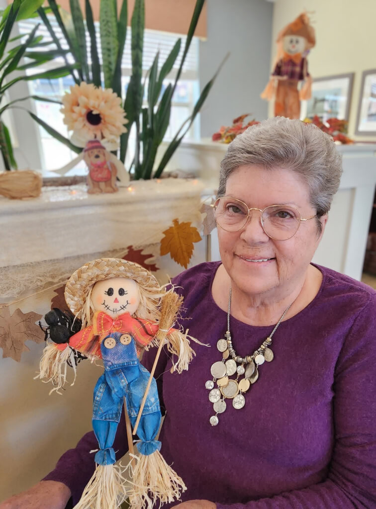 A smiling senior woman in a purple sweater holds a festive scarecrow decoration, surrounded by autumn garland and decorations at her community.