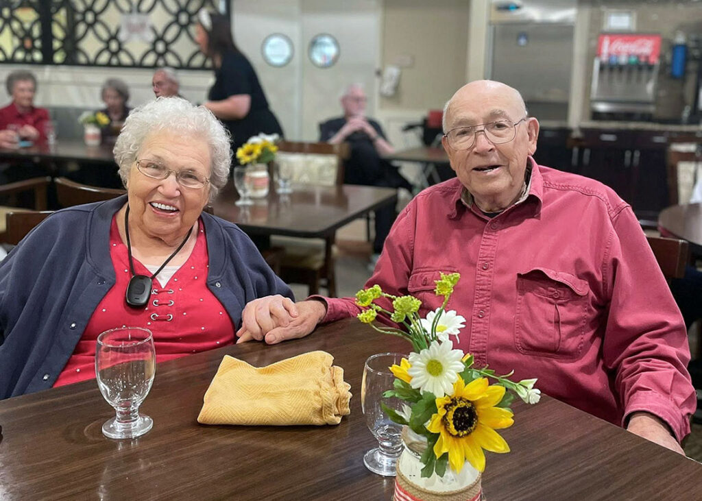 A senior man and woman hold hands at a dining table at The Wildwood, smiling with sunflowers in front of them.