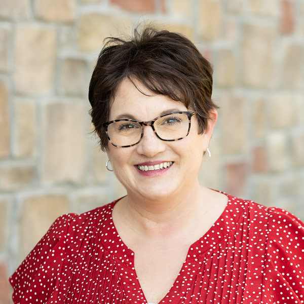 Becky Jacobs, Operations Coordinator at The Wildwood Senior Living, smiling in front of a tan stone wall. She has short dark hair, wears glasses, and is dressed in a red blouse with white polka dots.