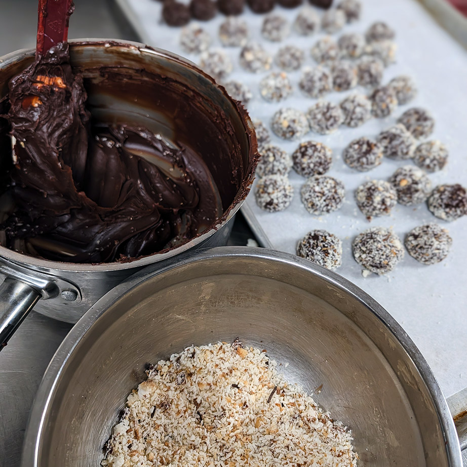 A pan of rich chocolate rests beside a sheet of freshly-made truffle balls.