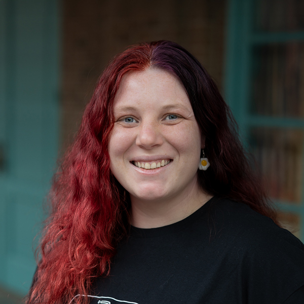 Karis Mengarelli, Culinary Director at The Wildwood Senior Living, smiling in a professional headshot, wearing a black top, with long red hair and a softly blurred brick and window background.