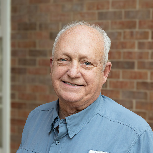 Mike Crafton, Plant Operations Director at The Wildwood Senior Living, smiling in a professional outdoor headshot with short white hair, a light blue button-up shirt, and a softly blurred brick background.