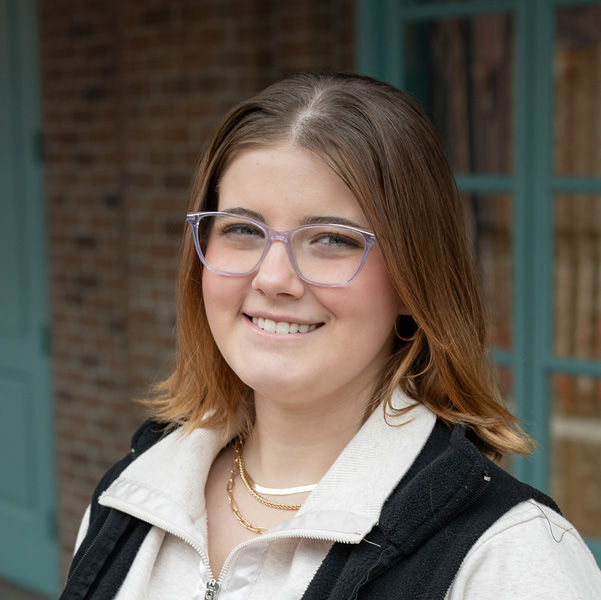 Rylee Sprague, Resident Services Director at The Wildwood Senior Living, smiling in a professional outdoor headshot with shoulder-length light brown hair, glasses, gold hoop earrings, and softly blurred brick and teal window details in the background.