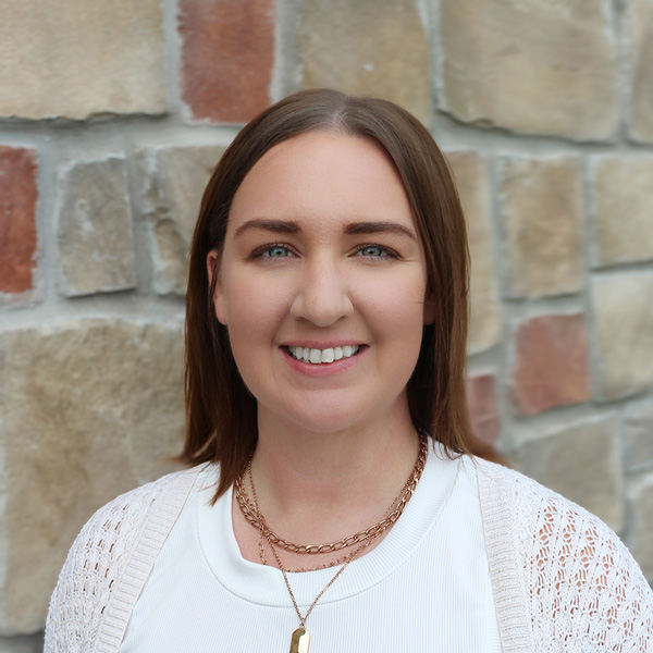 Stephanie Cormier, Senior Living Director at The Wildwood Senior Living, smiling in a professional headshot with straight brown hair, a white top, layered necklaces, and a stone wall background.