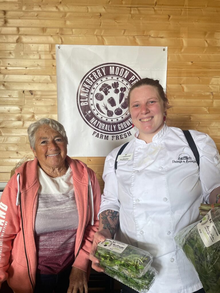 A Parker & Barrow staff member at The Wildwood Senior Living holds up fresh ingredients next to a senior resident while they both smile together.